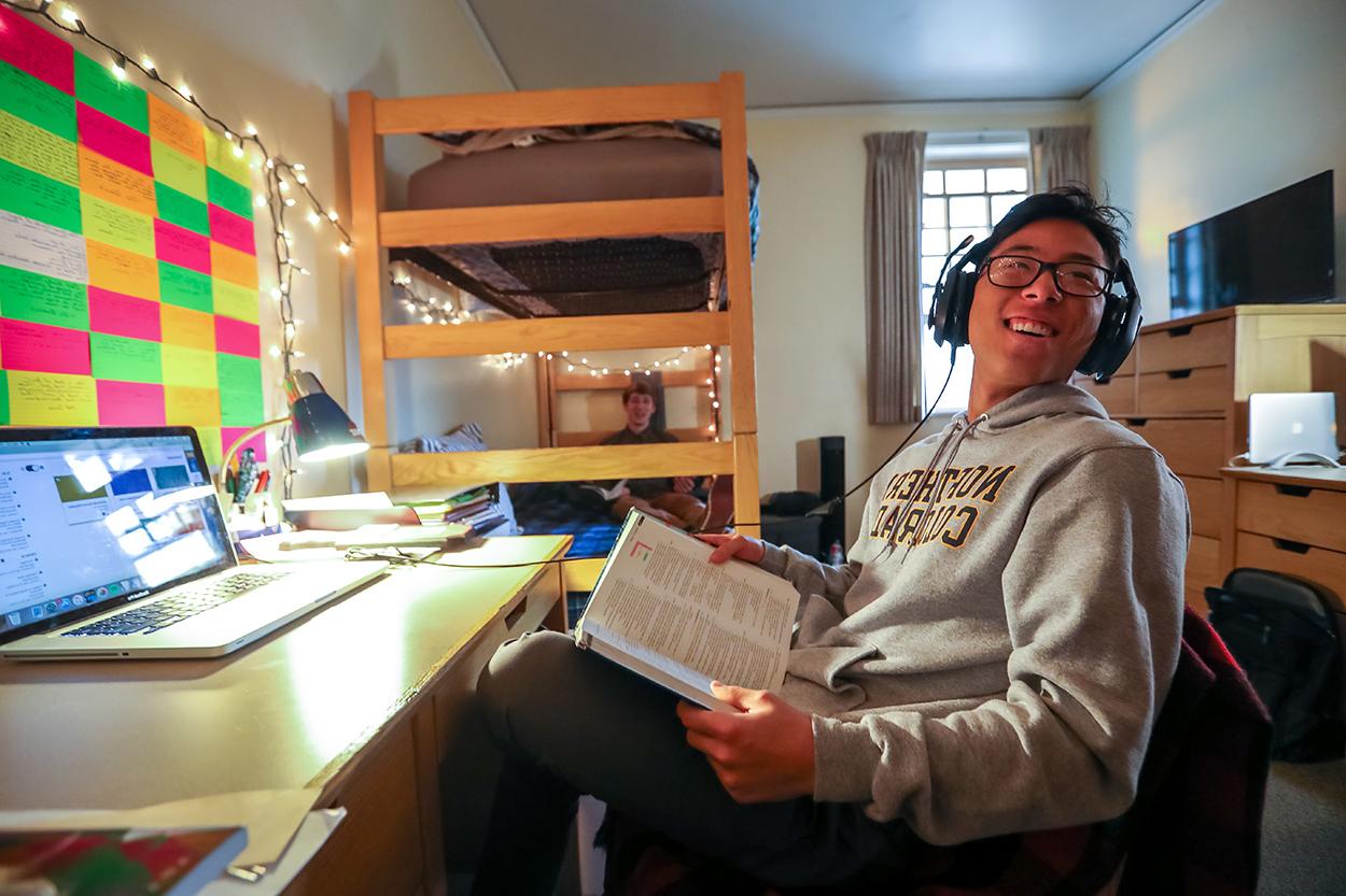 Student smiles while sitting at their desk holding a textbook in their new dorm room.